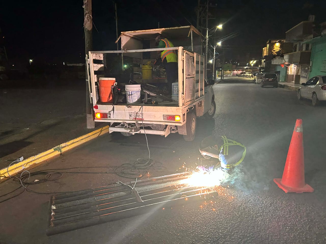 A worker is welding metal pipes on a street at night, with a truck and safety cones nearby.